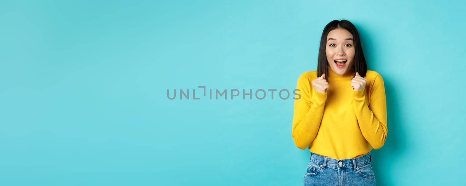 Hopeful and happy asian woman looking with amazement and hope at camera, winning, standing over blue background.