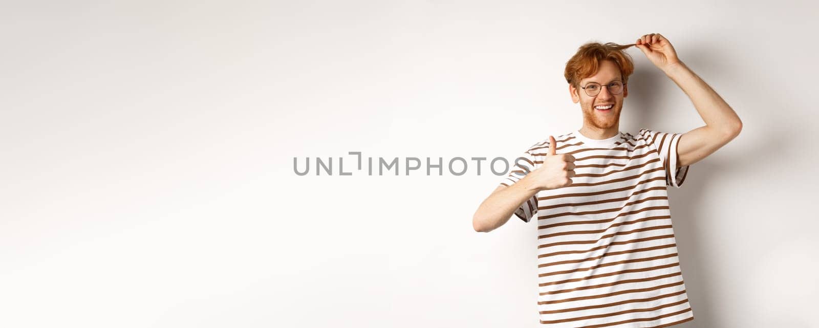 Cheerful redhead guy showing thumb-up and his red hair strand, like new color, standing over white background.