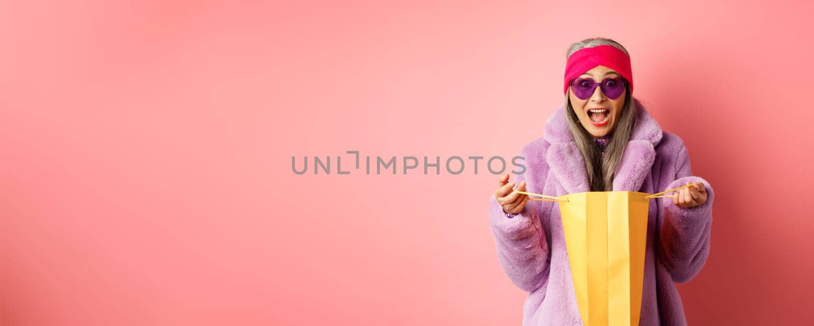 Shopping and fashion concept. Stylish asian elderly woman in sunglasses and faux fur coat open paper bag with gifts, looking amazed at camera, pink background.