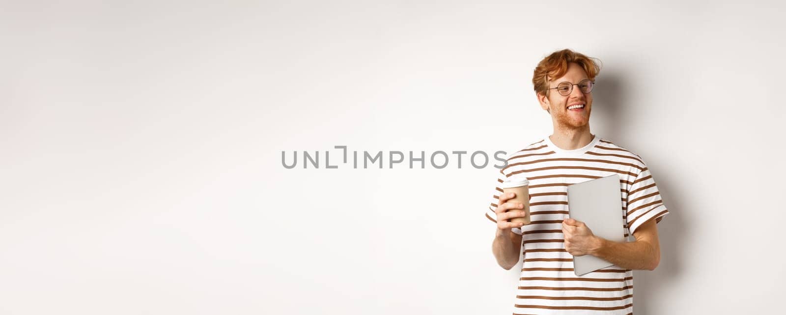 Handsome redhead male employee in glasses having break, drinking coffee and holding laptop, standing over white background.