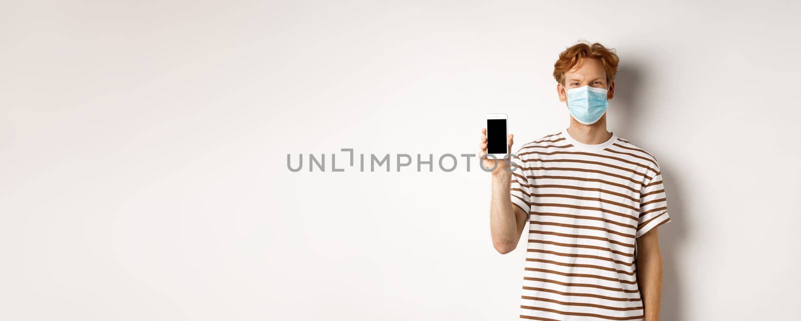 Covid-19, pandemic and social distancing concept. Handsome young man with face mask, showing black smartphone screen and smiling, standing over white background by Benzoix