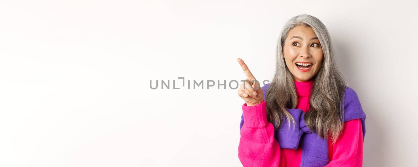 Close-up of stylish korean senior woman with grey hair looking and pointing upper left corner, showing promotion offer, standing over white background by Benzoix