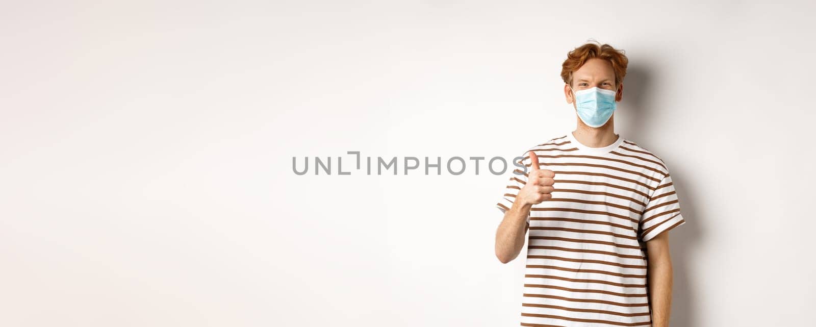 Covid-19, pandemic and social distancing concept. Young man with red hair wearing medical mask to prevent catching coronavirus, showing thumbs-up, white background by Benzoix