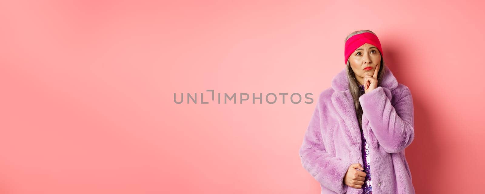 Funky old asian woman looking thoughtful and perplexed, standing in fake fur coat and thinking, making decision, standing on pink background.