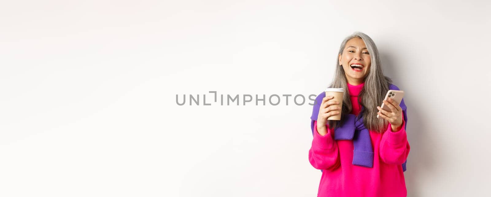 Social networking. Happy asian senior woman drinking coffee and holding smartphone, laughing at camera, standing over white background.