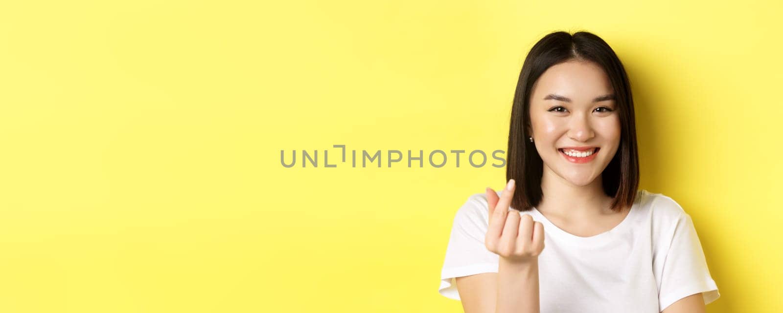 Valentines day and women concept. Close up of pretty asian girl in white t-shirt, smiling and showing finger heart, standing over yellow background.