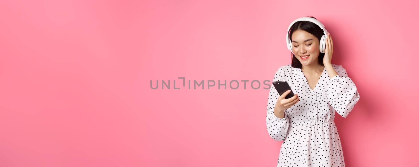 Cute asian girl listening music on headphones, looking at mobile phone and smiling, standing in dress against pink background by Benzoix