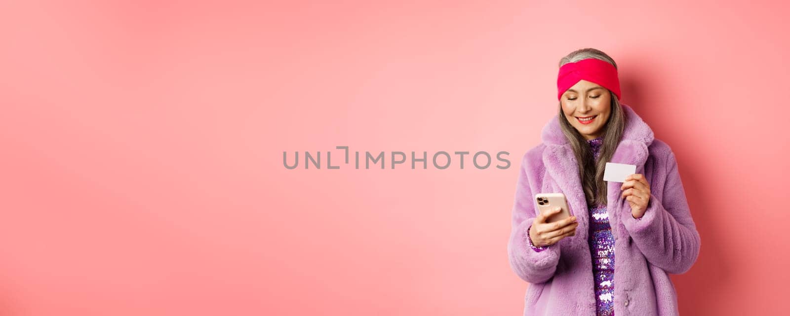 Online shopping and fashion concept. Stylish asian elderly woman standing in fashionable purple coat and making payment on smartphone, holding plastic credit card, pink background.