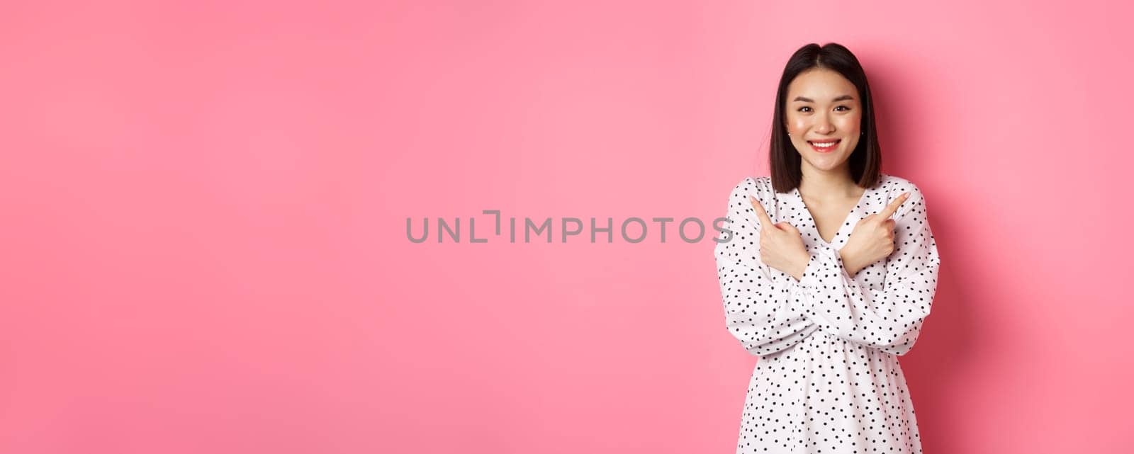 Beautiful asian girl in romantic dress pointing fingers sideways, showing two variants on shopping, smiling at camera, standing against pink background.