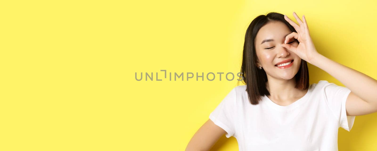 Close up of cute asian girl feeling happy, showing OK sign on eye and smiling, standing over yellow background by Benzoix