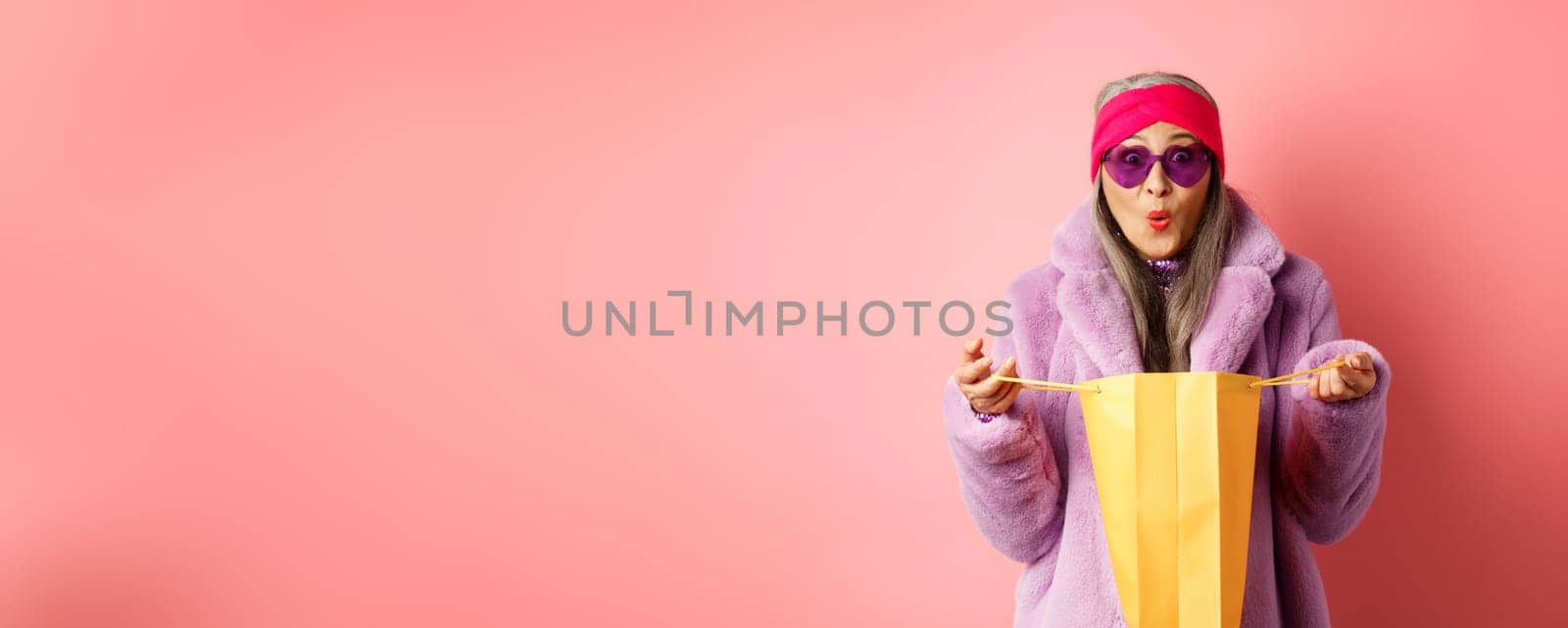 Shopping and fashion concept. Stylish asian elderly woman in sunglasses and faux fur coat open paper bag with gifts, looking surprised at camera, pink background.
