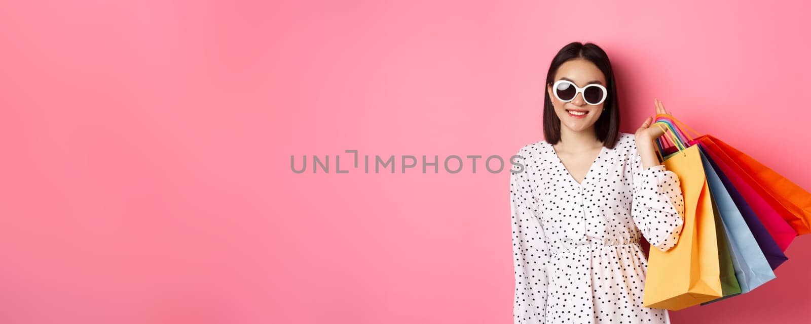 Young asian woman in sunglasses going shopping, holding bags from malls and stores and smiling, standing over pink background by Benzoix