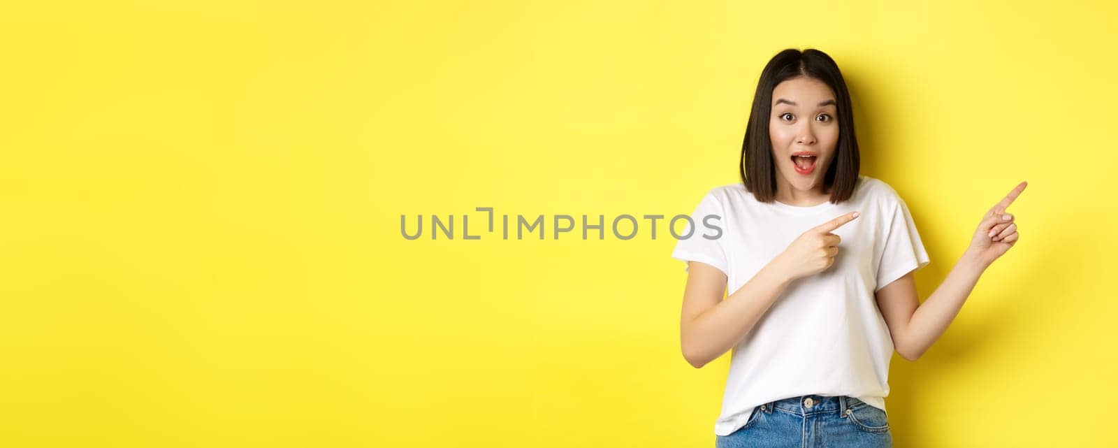 Beauty and fashion concept. Beautiful asian woman in white t-shirt pointing fingers right, demonstrate logo standing over yellow background by Benzoix