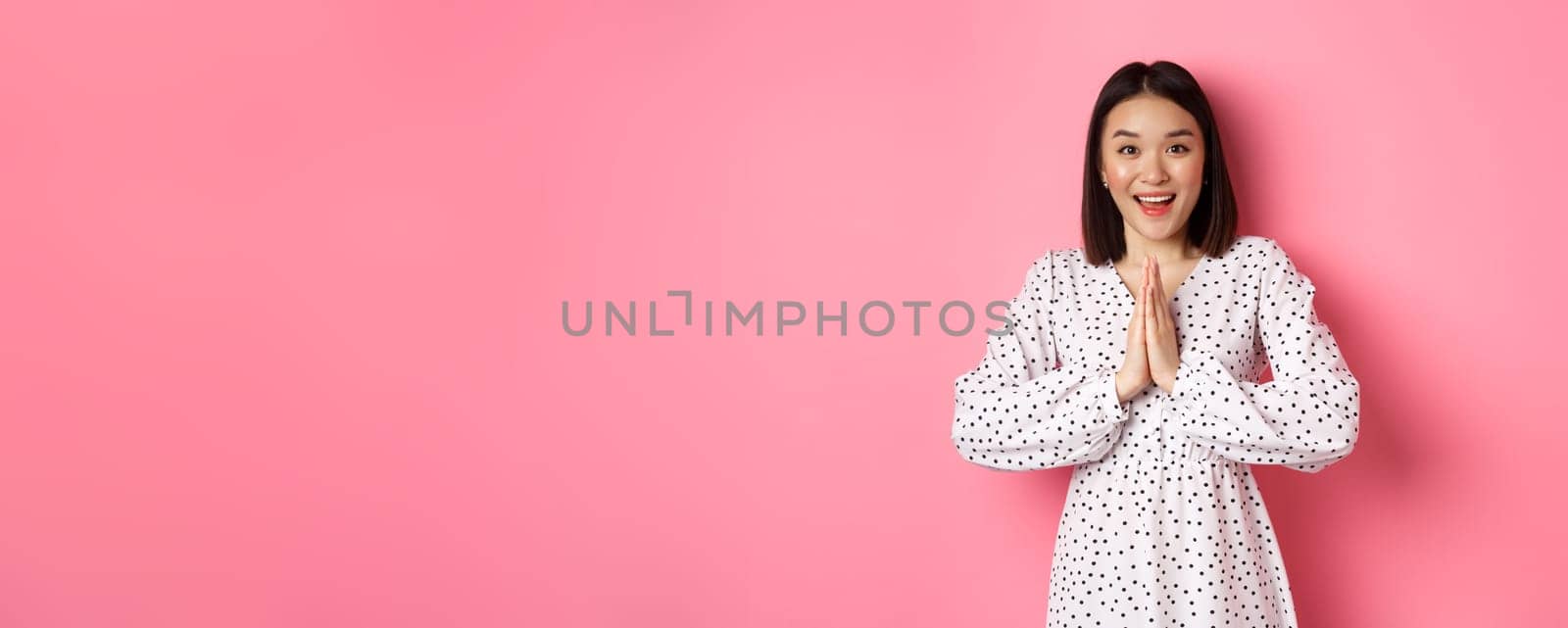 Beautiful asian woman thanking you, holding hands together in appreciation gesture, smiling happy at camera, standing grateful over pink background.