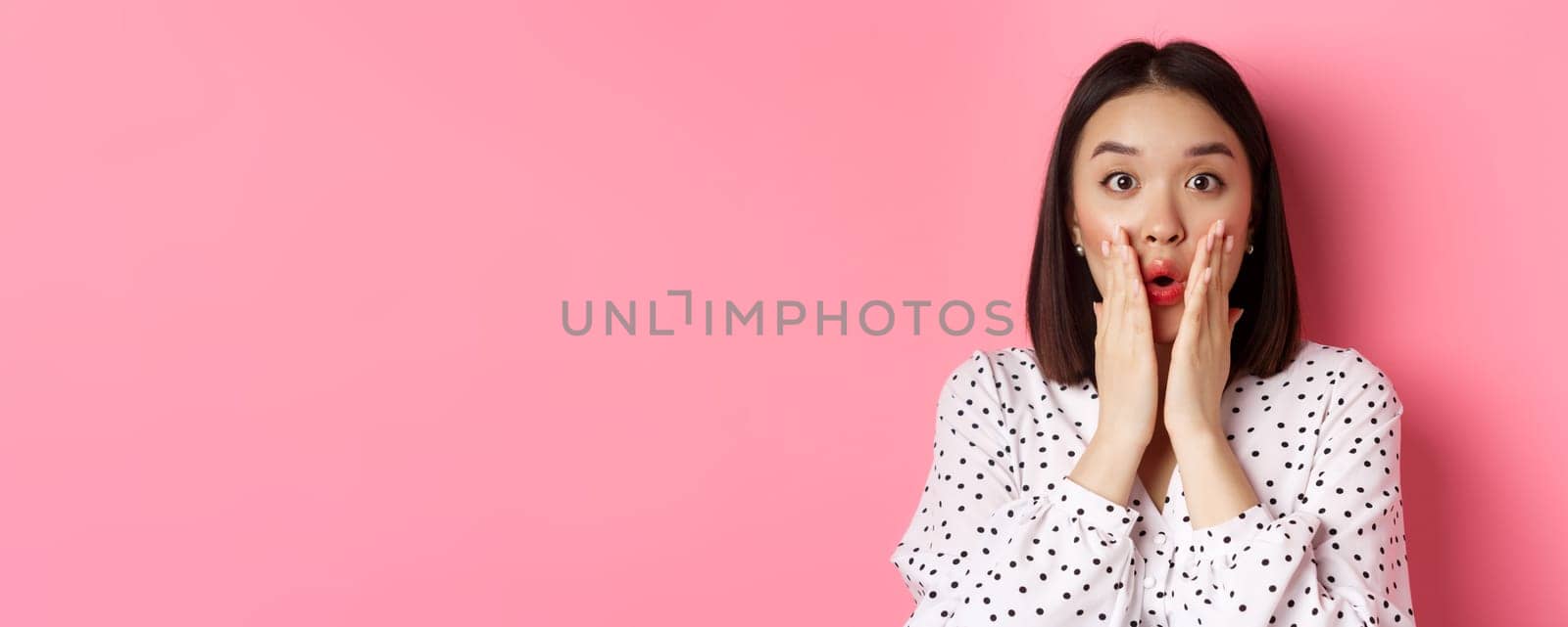 Close-up of surprised and amazed asian woman staring at camera, gasping impressed, holding hands near mouth, standing over pink background by Benzoix
