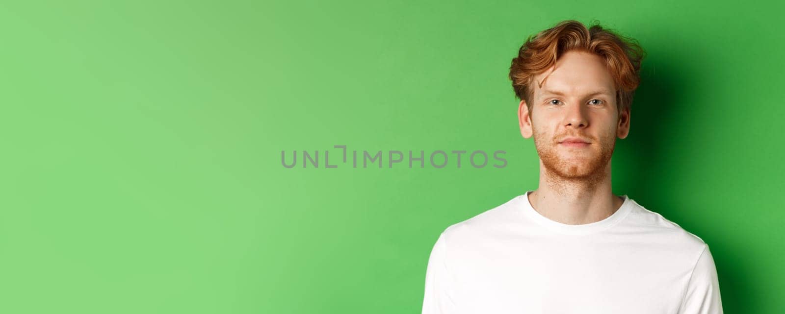 Close-up of young man with red messy hair and beard looking at camera, standing over green background.