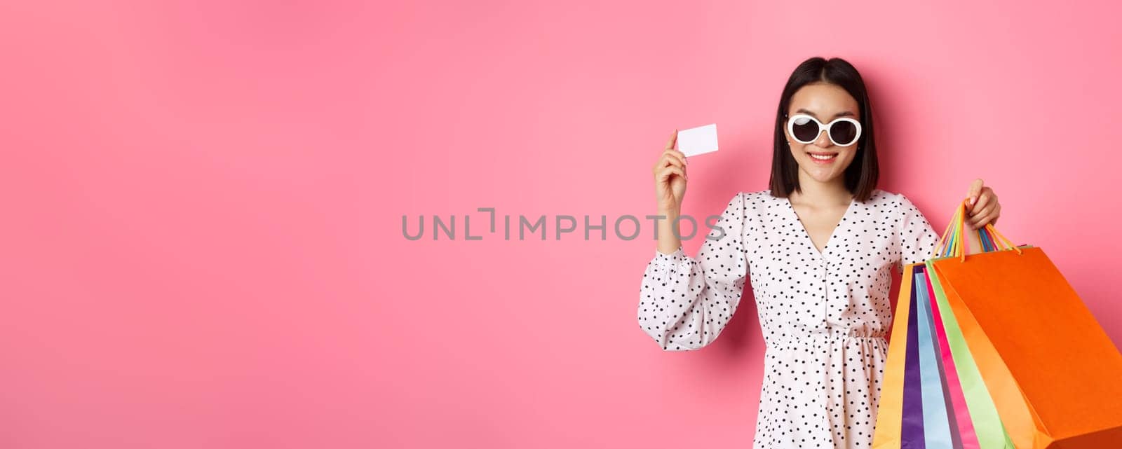 Beautiful asian woman in sunglasses going shopping, holding bags and showing credit card, standing over pink background by Benzoix
