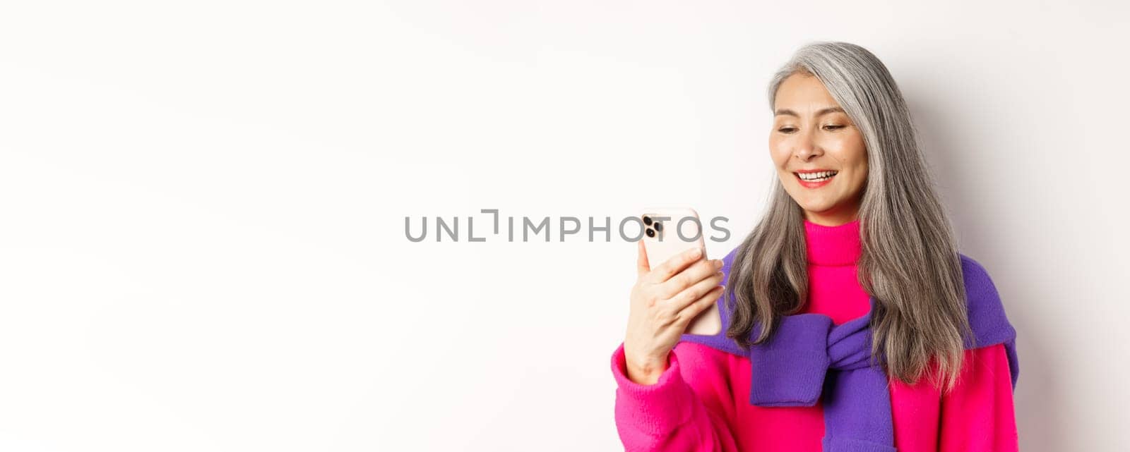 Close-up of fashionable asian senior woman looking at smartphone and reading message, smiling at mobile screen, standing over white background by Benzoix