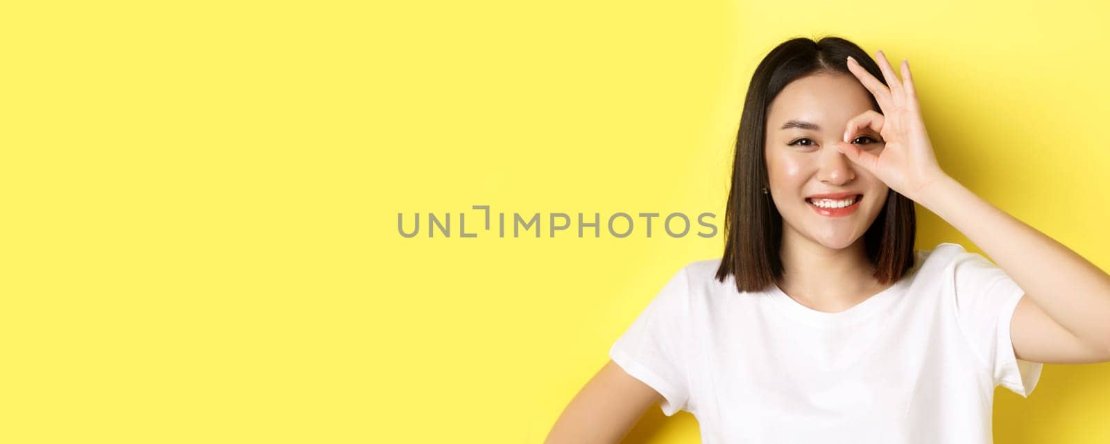 Close up of cute asian girl feeling happy, showing OK sign on eye and smiling, standing over yellow background.