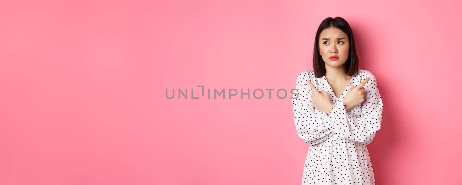 Concerned asian woman having doubts, pointing sideways and looking left with hesitant and sad face, need help with choice, standing over pink background by Benzoix