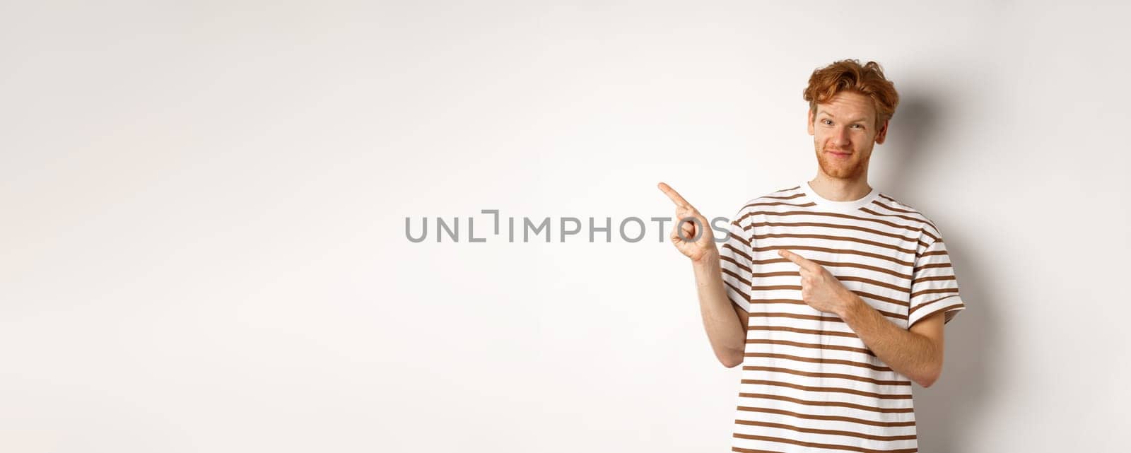 Doubtful redhead guy pointing fingers left at looking skeptical at camera, standing over white background by Benzoix