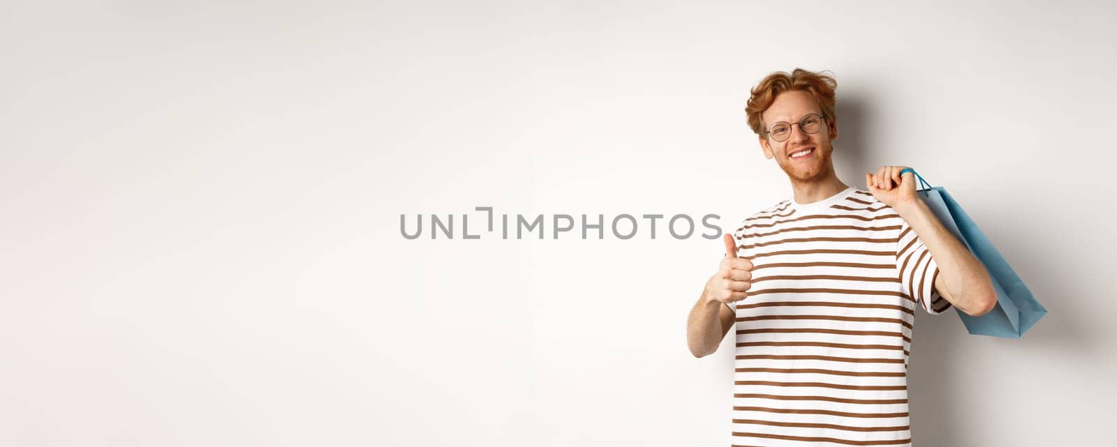 Satisfied young man leave positive review after shopping, showing thumb-up in approval and smiling, holding paper bag over shoulder, white background.