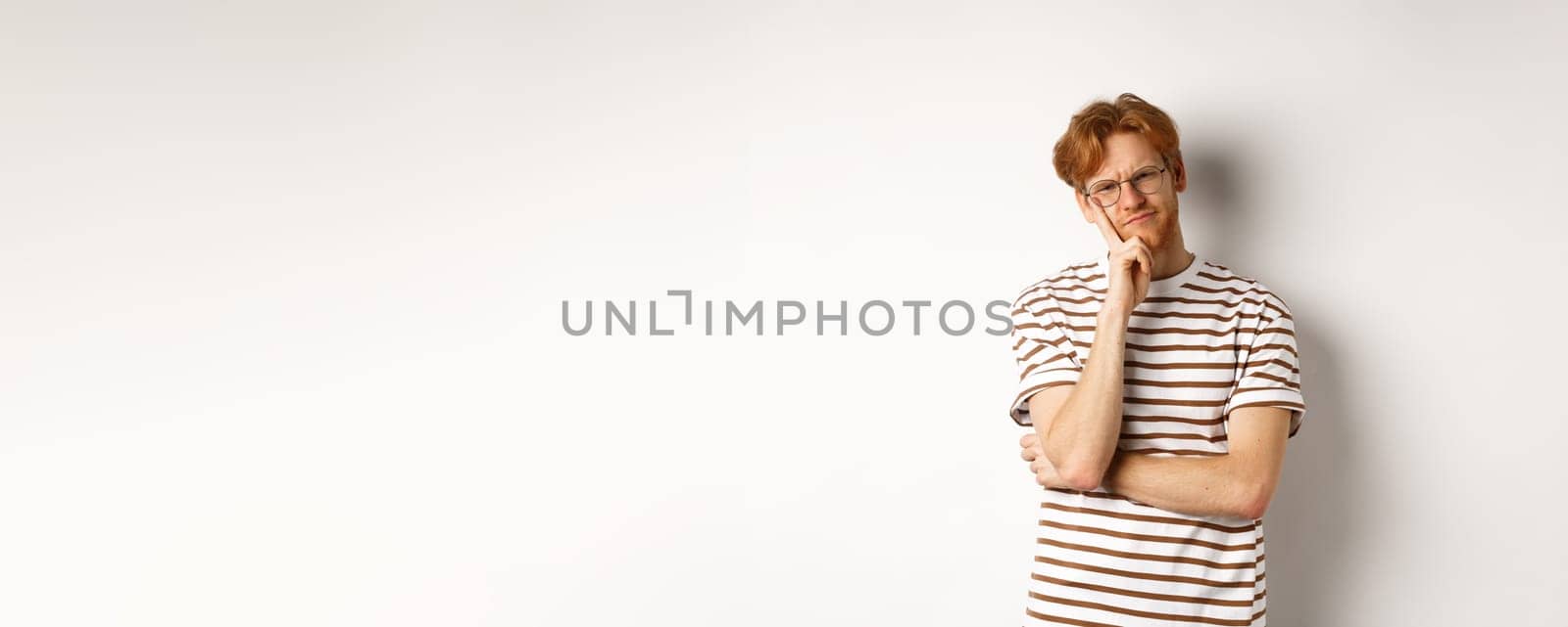 Young man with red messy hair and glasses looking doubtful, thinking and squinting at camera, standing thoughtful over white background by Benzoix