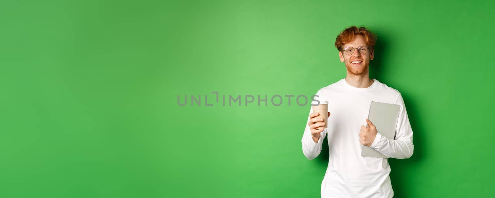 Handsome young man, employee with laptop drinking coffee on break, smiling satisfied, standing over green background.