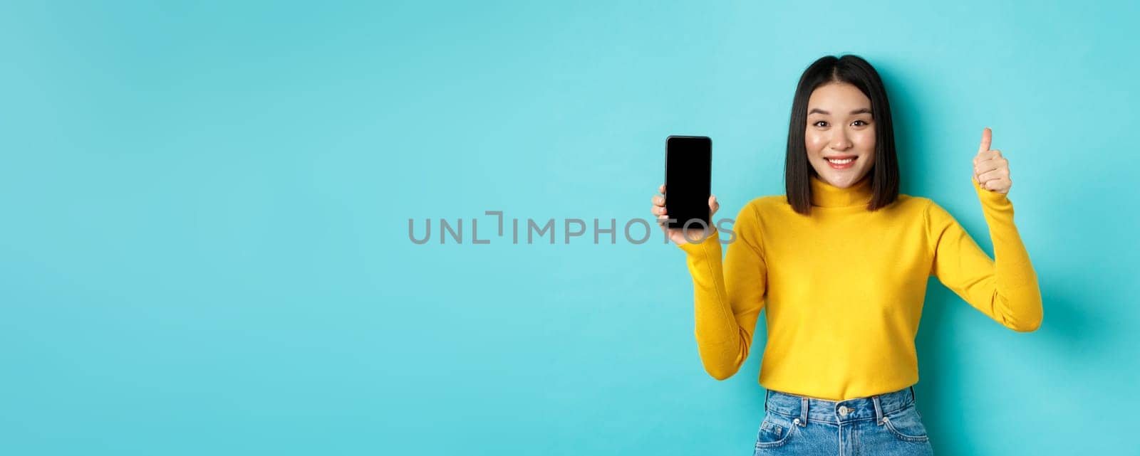 Technology and people concept. Cheerful asian girl in yellow sweater showing blank smartphone screen and thumbs up, demonstrate online offer, standing over blue background.