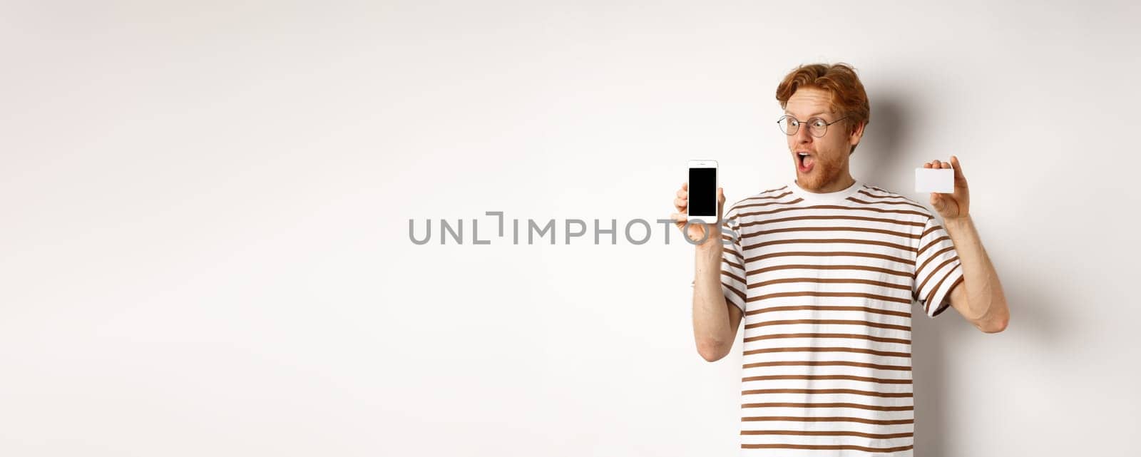 Shopping and finance concept. Amazed young man with red hair showing plastic credit card and smartphone blank screen, staring at display impressed, white background by Benzoix