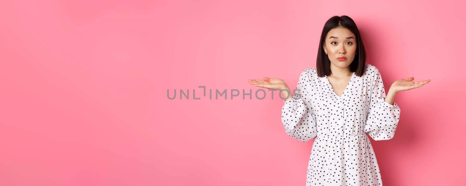 Indecisive cute asian woman shrugging, holding hands spread sideways on copy spaces, dont know, having doubts, standing over pink background by Benzoix