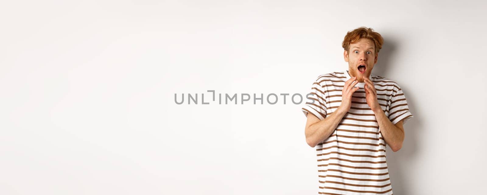 Timid young man with red hair, looking scared, jumping and screaming from fear, standing over white background.