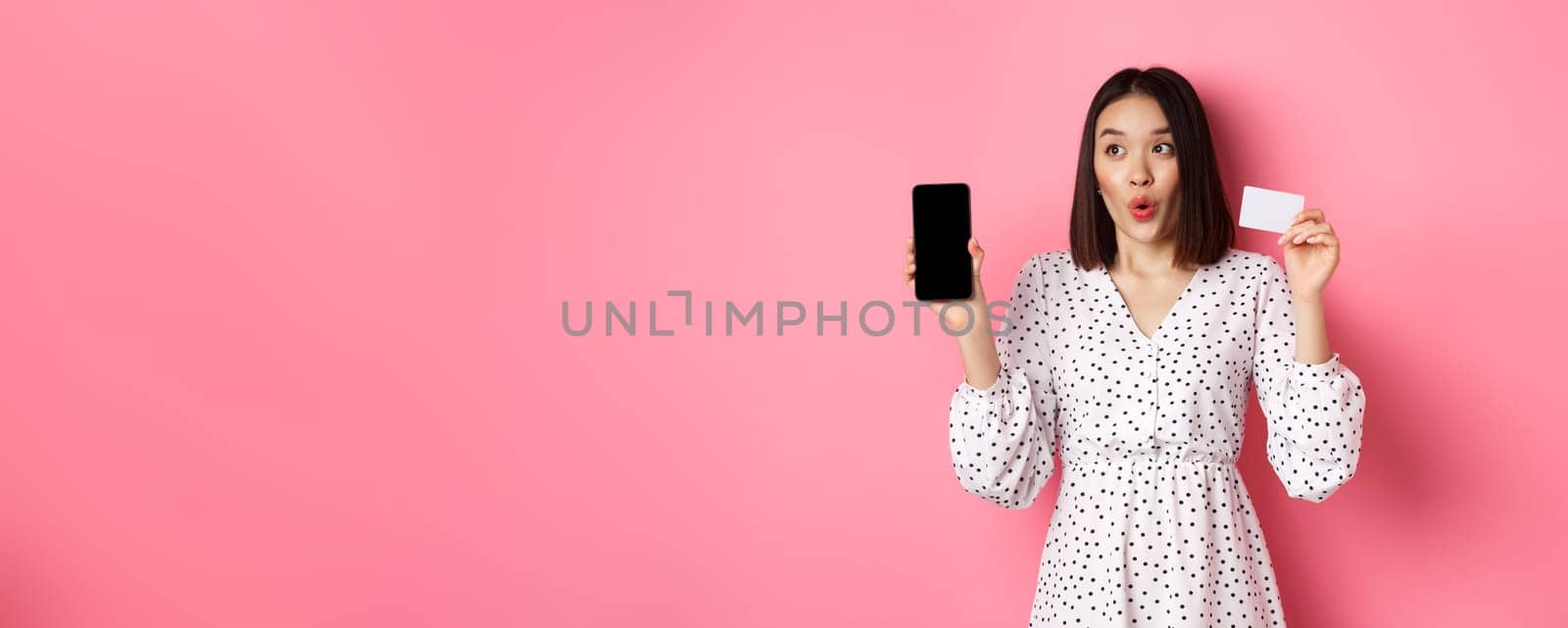 Cute asian woman shopping online, showing bank credit card and mobile screen, smiling and looking left at copy space, standing over pink background.