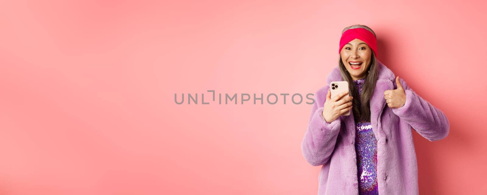 Online shopping and fashion concept. Happy asian grandmother checking out awesome promo, pointing finger at smartphone and looking amazed, pink background.