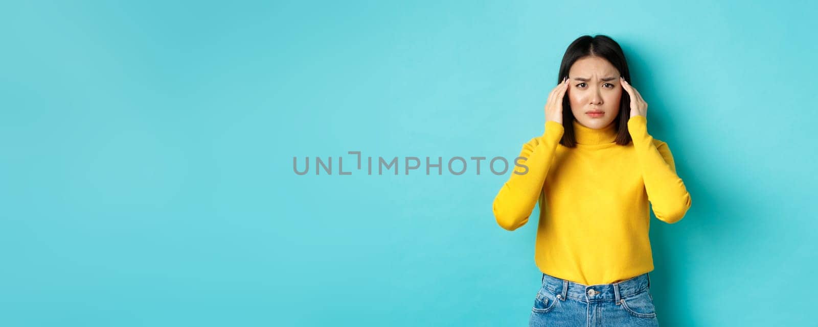 Image of distressed asian woman touching head and frowning, feeling headache, standing troubled against blue background.