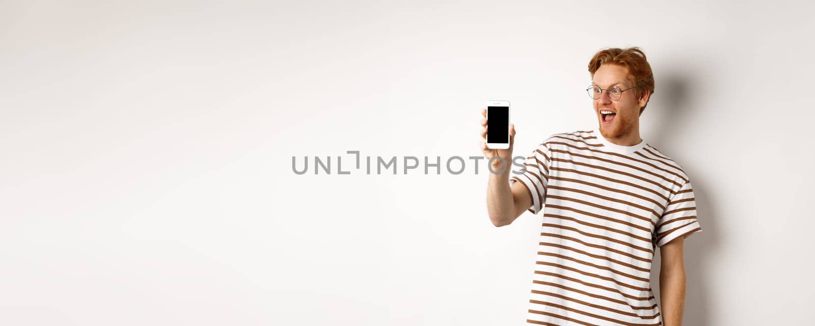 Technology and e-commerce concept. Happy young redhead man in glasses showing blank smartphone screen, looking amazed, standing over white background by Benzoix