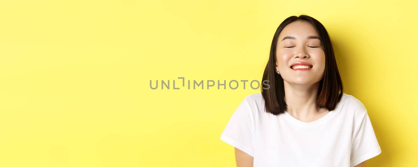 Close up of happy and relaxed asian woman enjoying sun, smiling with eyes closed and looking joyful, standing over yellow background.