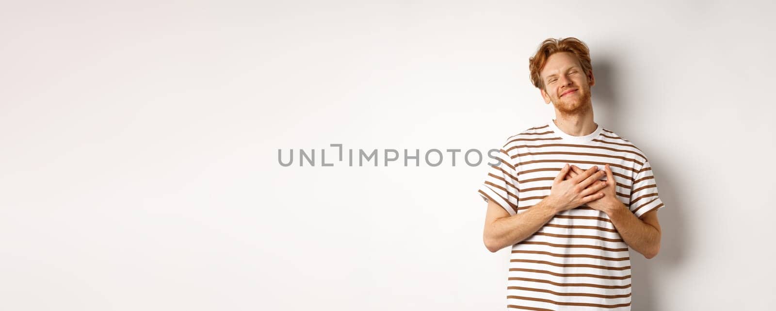 Happy young man with ginger hair appreciate gift, holding hands on heart and smiling, thanking you, standing grateful against white background.