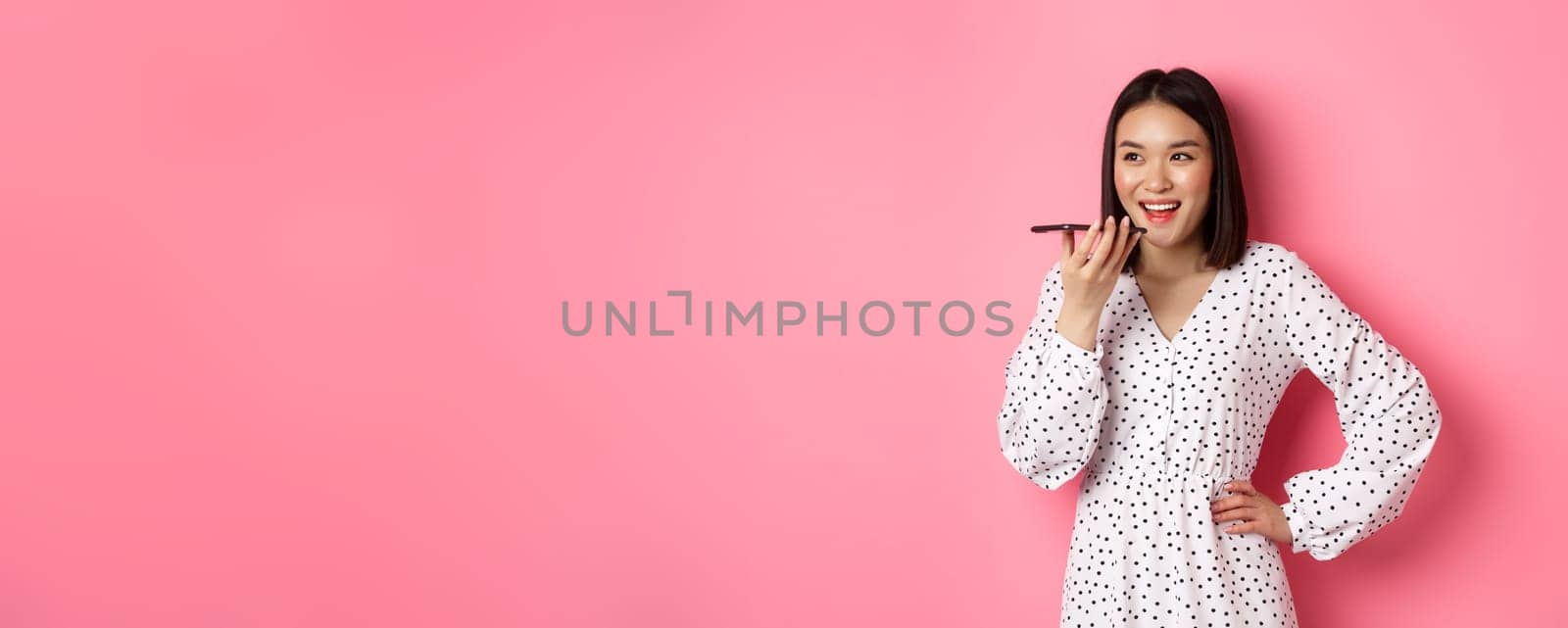 Beautiful korean woman talking on speakerphone, recording voice message and smiling happy, standing over pink background.