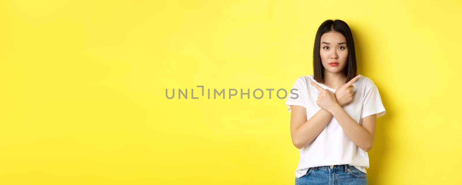 Indecisive asian girl need help with choice, pointing fingers sideways and looking confused, standing over yellow background by Benzoix