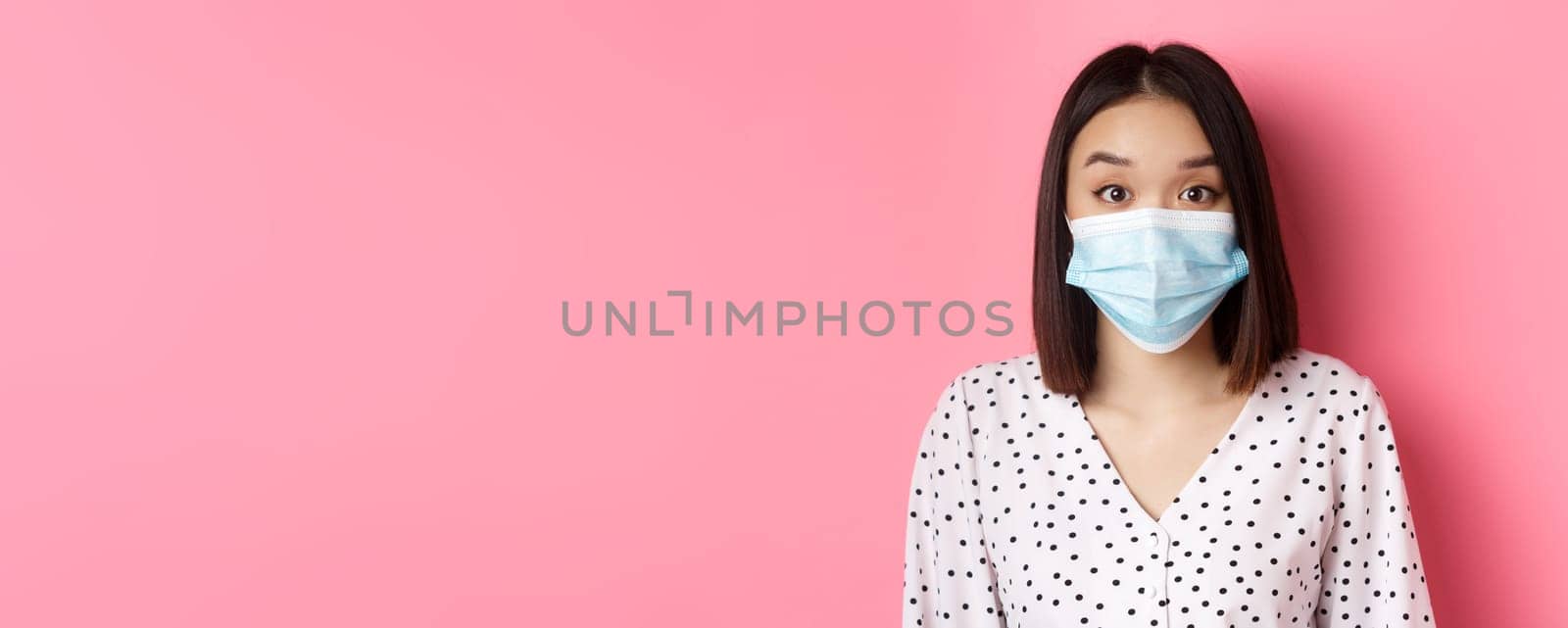 Covid-19, pandemic and lifestyle concept. Surprised asian woman in face mask raising eyebrows, staring at camera amazed, standing against pink background by Benzoix