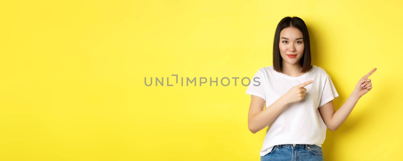 Beauty and fashion concept. Beautiful asian woman in white t-shirt pointing fingers right, demonstrate logo standing over yellow background by Benzoix