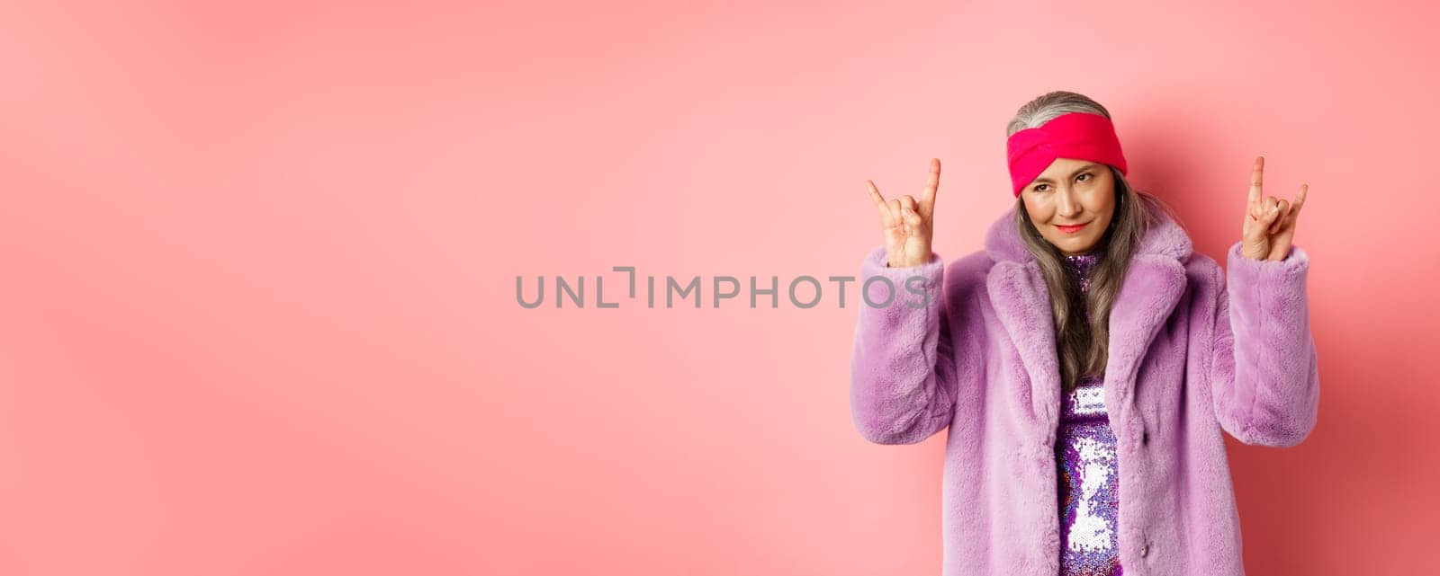Funny and cool asian senior woman showing rock n roll gesture, looking sassy, standing over pink background.