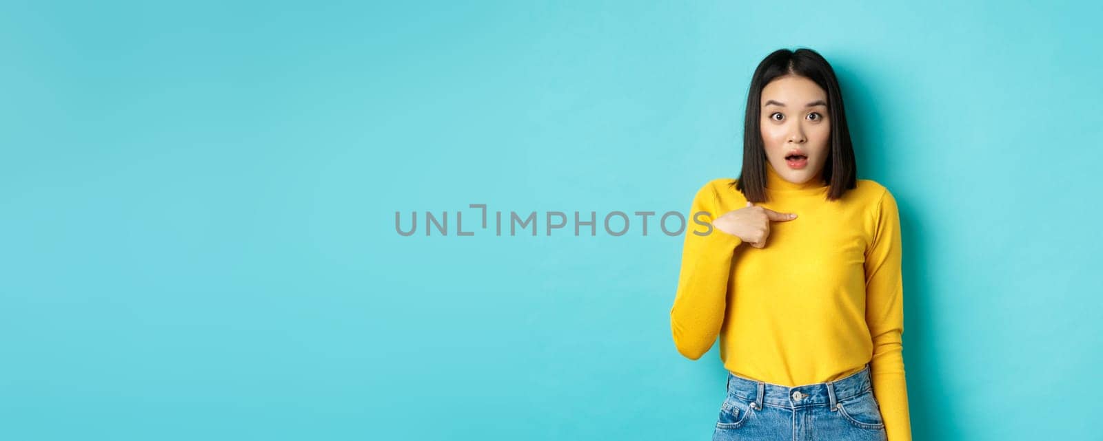 Image of surprised asian girl gasping and pointing at herself with disbelief, standing over blue background.