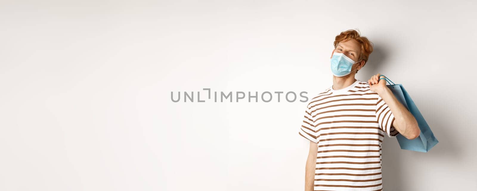 Concept of covid-19 and shopping. Satisfied young man looking pleased after shopping, wearing face mask, holding paper bag and smiling, white background.