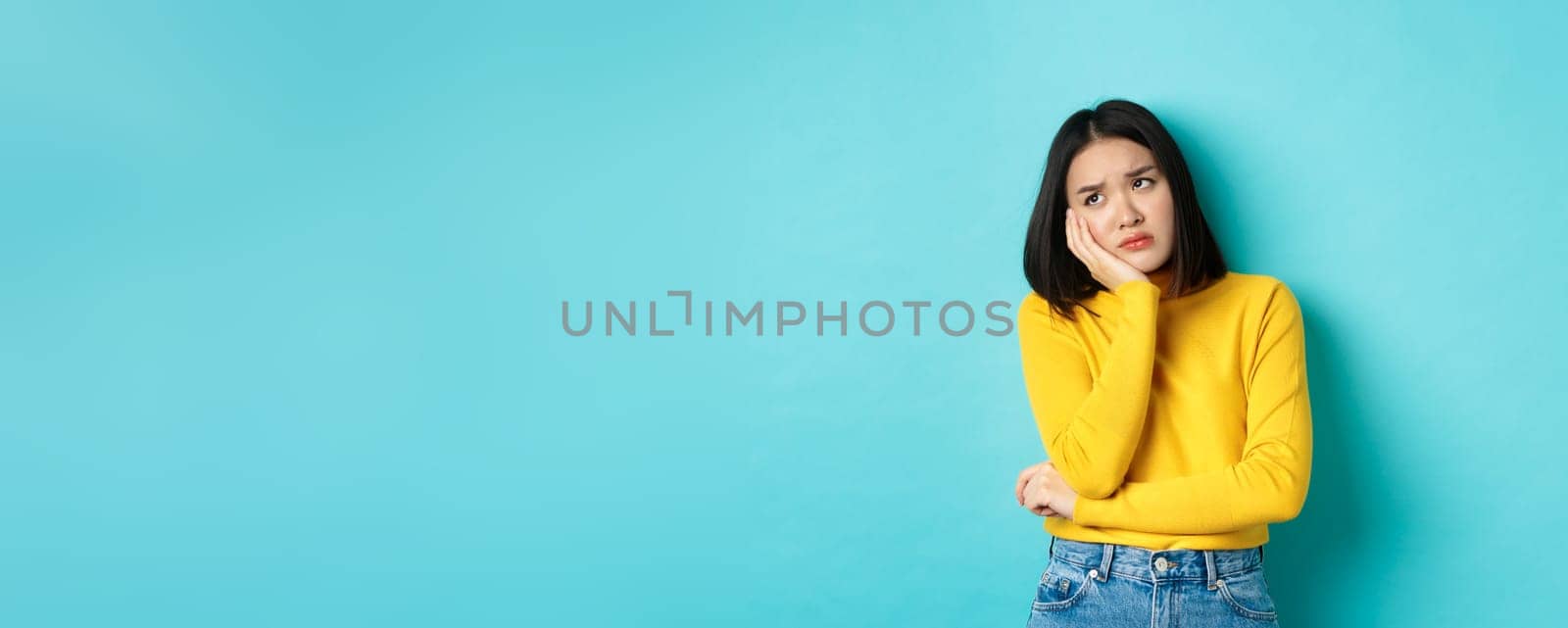 Sad and bored asian teenage girl leaning on palm, looking at upper left corner with lonely and upset expression, standing over blue background.