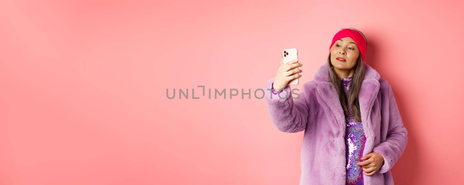 Fashion concept. Stylish asian senior female taking selfie on smartphone, posing in purple faux fur coat and party dress, standing over pink background by Benzoix