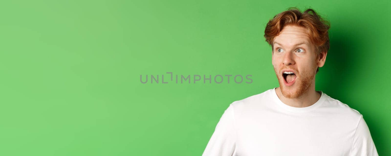 Close up of surprised and impressed redhead man checking out promotion offer, looking left with dropped jaw, standing over green background by Benzoix