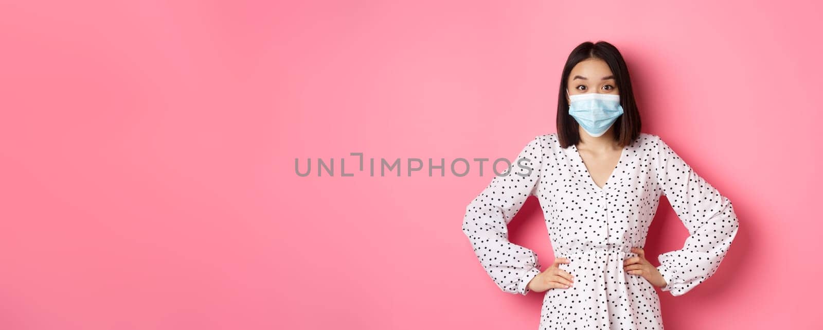 Covid-19, quarantine and lifestyle concept. Cute korean woman in dress and face mask using preventive measures from coronavirus, standing over pink background by Benzoix
