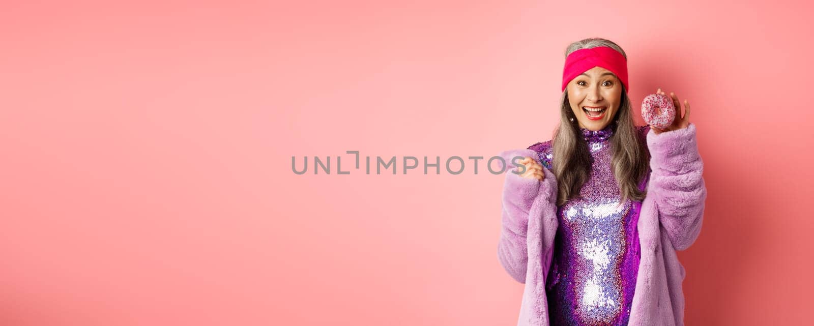 Cheerful asian senior woman with grey hair, wearing party glitter dress, showing delicious donut and smiling happy, eating sweet food, standing over pink background by Benzoix